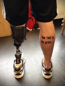 Фото тату надписи "One foot in the grave..." на ноге мужчины с протезом. Смелый и дерзкий рисунок татуировки.