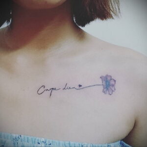 Фото тату надписи "Carpe Diem" с небольшим акварельным цветком на ключице девушки. Нежный и вдохновляющий рисунок татуировки.