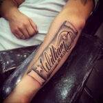 Фото тату: черная татуировка с именем 'William' и датой '02.10.13' на ленте, украшенной звездами, на предплечье мужчины.