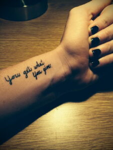 Фото тату надписи 'You get what you give' на запястье. Черный цвет, каллиграфический шрифт, минималистичный дизайн.