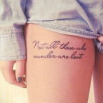 Фото тату: изящная надпись 'Not all those who wander are lost' на бедре девушки, выполненная тонким черным контуром.