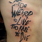 Фото тату: черная надпись 'Too Weird to Live Too Rare to Die' на боку человека, выполненная в стиле леттеринга с добавлением силуэтов летучих мышей.