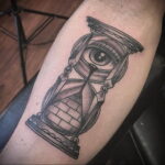 Фото тату: татуировка в стиле blackwork с изображением глаза внутри песочных часов, выполненная на руке мужчины.