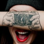 Фотография татуировки на руке, изображающей старинную камеру Pentax. Реалистичный рисунок с детальной проработкой оптики и корпуса.