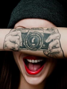 Фотография татуировки на руке, изображающей старинную камеру Pentax. Реалистичный рисунок с детальной проработкой оптики и корпуса.