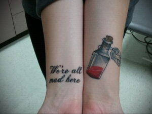 Фото тату с цитатой 'We're all mad here' и изображением бутылочки с надписью 'Drink Me'. Татуировка на лодыжках в стиле иллюстрации.