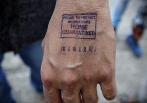Фотография руки с синим штампом карантина, на котором написано 'Home Quarantined'. Изображение демонстрирует временную татуировку.