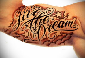 Фото тату на руке: надпись "Give the Dream" в каллиграфическом стиле, окруженная звездами и облаками. Черно-коричневая гамма.