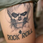 Фото тату: татуировка на ноге с изображением черепа в стиле гранж и надписью "ROCK 'N' ROLL". Черно-белая татуировка с агрессивным дизайном.