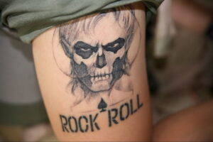 Фото тату: татуировка на ноге с изображением черепа в стиле гранж и надписью "ROCK 'N' ROLL". Черно-белая татуировка с агрессивным дизайном.
