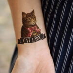 Фото татуировки с изображением милого кота в бантике и надписью "Cat Lady" на женском запястье. Яркий рисунок татуировки выполнен в стиле олдскул.