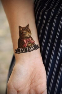Фото татуировки с изображением милого кота в бантике и надписью "Cat Lady" на женском запястье. Яркий рисунок татуировки выполнен в стиле олдскул.