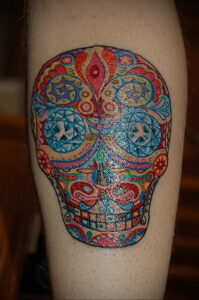 Фото тату черепа в стиле Sugar Skull на икре. Многоцветный рисунок с детальной прорисовкой узоров и орнаментов. Отличный эскиз тату для ценителей мексиканской культуры.