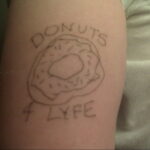 Фото тату на руке: простой рисунок пончика с надписью 'DONUTS 4 LYFE', выполненный в минималистичном стиле черными линиями.