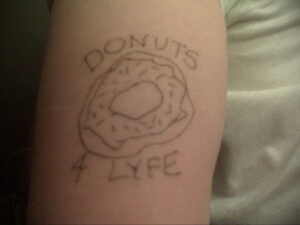 Фото тату на руке: простой рисунок пончика с надписью 'DONUTS 4 LYFE', выполненный в минималистичном стиле черными линиями.