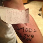Фото татуировки на руке с надписью "Daddy I love you" и рисунком детской фигурки. Черно-белый рисунок, выполненный от руки.