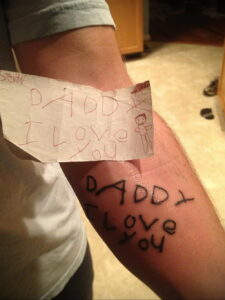 Фото татуировки на руке с надписью "Daddy I love you" и рисунком детской фигурки. Черно-белый рисунок, выполненный от руки.