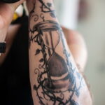 Фото тату на руке: песочные часы, обвитые цветами и ветвями, выполненные в черно-серых тонах.