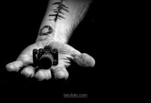 Черно-белое фото руки, держащей миниатюрную камеру Fujifilm. На предплечье видны татуировки в виде геометрических фигур и абстрактных узоров. Символизирует любовь к фотографии.