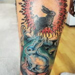 Фото тату зайцев на ноге, выполненное в ярких цветах с использованием красного, синего и зеленого. Детализированный рисунок с элементами пламени и растительности.