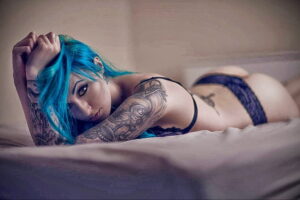 Фото девушки с яркими голубыми волосами и множеством татуировок в стиле дарк-арт на руках и теле. Изображение выполнено в темных тонах.