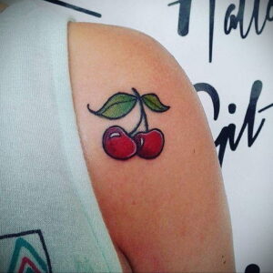 Фото тату: Красочная татуировка двух вишен с зелеными листьями на плече. Рисунок выполнен в стиле олдскул с использованием ярких цветов и четких контуров.