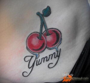 Фото тату двух вишен с капающими каплями и надписью "Yummy" на животе. Яркий и игривый рисунок в стиле олдскул.