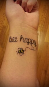 Фото татуировки с надписью "bee happy" и маленькой пчелой на запястье. Черно-белый рисунок татуировки с каллиграфической надписью и простой формой пчелы.