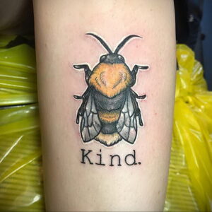 Фото тату пчелы на руке с надписью 'Kind'. Детализированный рисунок татуировки с реалистичными крыльями и телом.