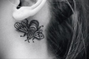 Фото минималистичной татуировки шмеля за ухом, выполненной в технике blackwork. Четкие линии и лаконичный дизайн.