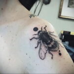 Фото тату шмеля на плече, выполненное в технике дотворк с использованием черных точек и линий. Рядом с насекомым расположены небольшие черные точки.
