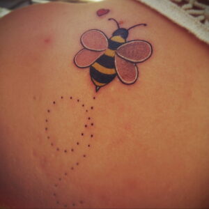 Фото татуировки пчелы, летящей к сердечку, выполненной в пастельных тонах. Рисунок расположен на спине.