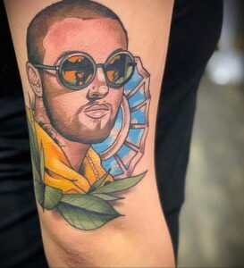 Фотография цветной татуировки на руке с изображением Mac Miller в солнцезащитных очках и с абстрактным сине-желтым фоном.