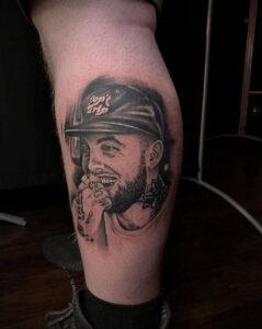 Фотография цветной татуировки на ноге с изображением Mac Miller в кепке, с сигаретой и улыбкой.