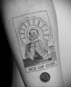 Черно-белое фото татуировки на руке с изображением Mac Miller в стиле карты Таро 'Ace of Cups'.