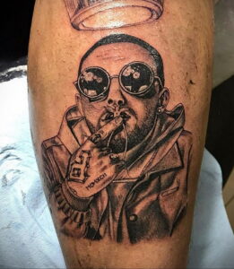 Черно-белое фото татуировки на руке с изображением Mac Miller в солнцезащитных очках, с сигаретой и короной.