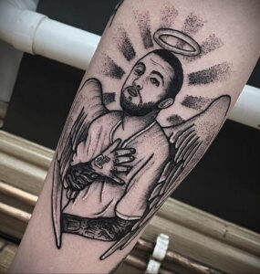 Черно-белое фото татуировки на предплечье с изображением портрета Mac Miller в образе ангела с нимбом и крыльями.
