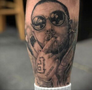 Черно-белое фото тату на предплечье с реалистичным изображением Mac Miller в очках и с сигаретой, с детально проработанными чертами лица.