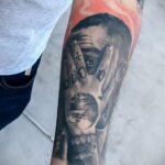 Черно-белое фото тату на предплечье с портретом Mac Miller, закрывающим лицо руками, с пламенем на фоне, символизирующим боль и страдание.