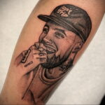 Черно-белое фото тату на предплечье с изображением Mac Miller в кепке, с сигаретой в руке и цветком рядом.