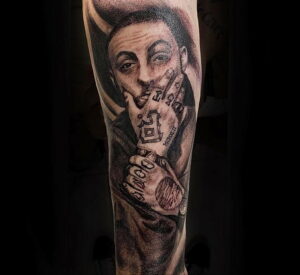 Черно-белое фото тату на предплечье с портретом Mac Miller, татуировками на пальцах, кольцами и датой MCMXCII.