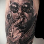 Фото тату на руке с реалистичным портретом Mac Miller в очках и с сигаретой. Детализированный рисунок татуировки.