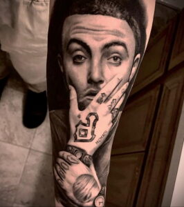 Фото тату на руке с крупным портретом Mac Miller, показывающего поднятый палец. Детализированный рисунок татуировки.