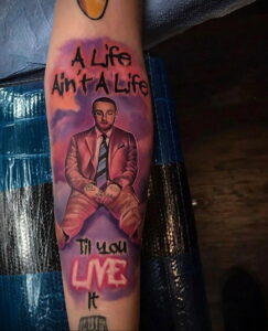 Фото тату на руке с изображением Mac Miller в костюме и с цитатой "A Life Ain't A Life Til You Live It". Яркий и детализированный рисунок татуировки.