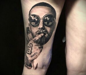 Фото тату на руке с реалистичным черно-белым портретом Mac Miller в круглых очках и с сигарой. Детализированный рисунок татуировки.