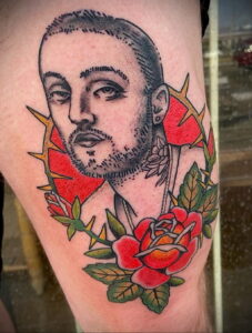 Фото тату на руке с реалистичным портретом Mac Miller, окруженного красной колючей проволокой и красными розами. Детализированный рисунок татуировки.