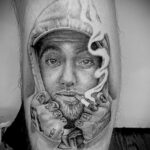 Черно-серая реалистичная татуировка Mac Miller с сигаретой во рту и кепкой на голове на руке.