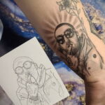 Цветная татуировка Mac Miller в скафандре космонавта на руке, с эскизом рядом и элементами цветочного фона.