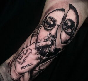 Детализированная черно-серая татуировка портрета Mac Miller на руке, с элементами символики и римскими цифрами.