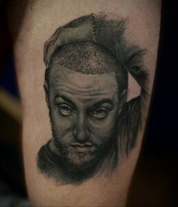 Черно-серая реалистичная татуировка портрета Mac Miller на предплечье. Детализированное изображение лица и кепки.